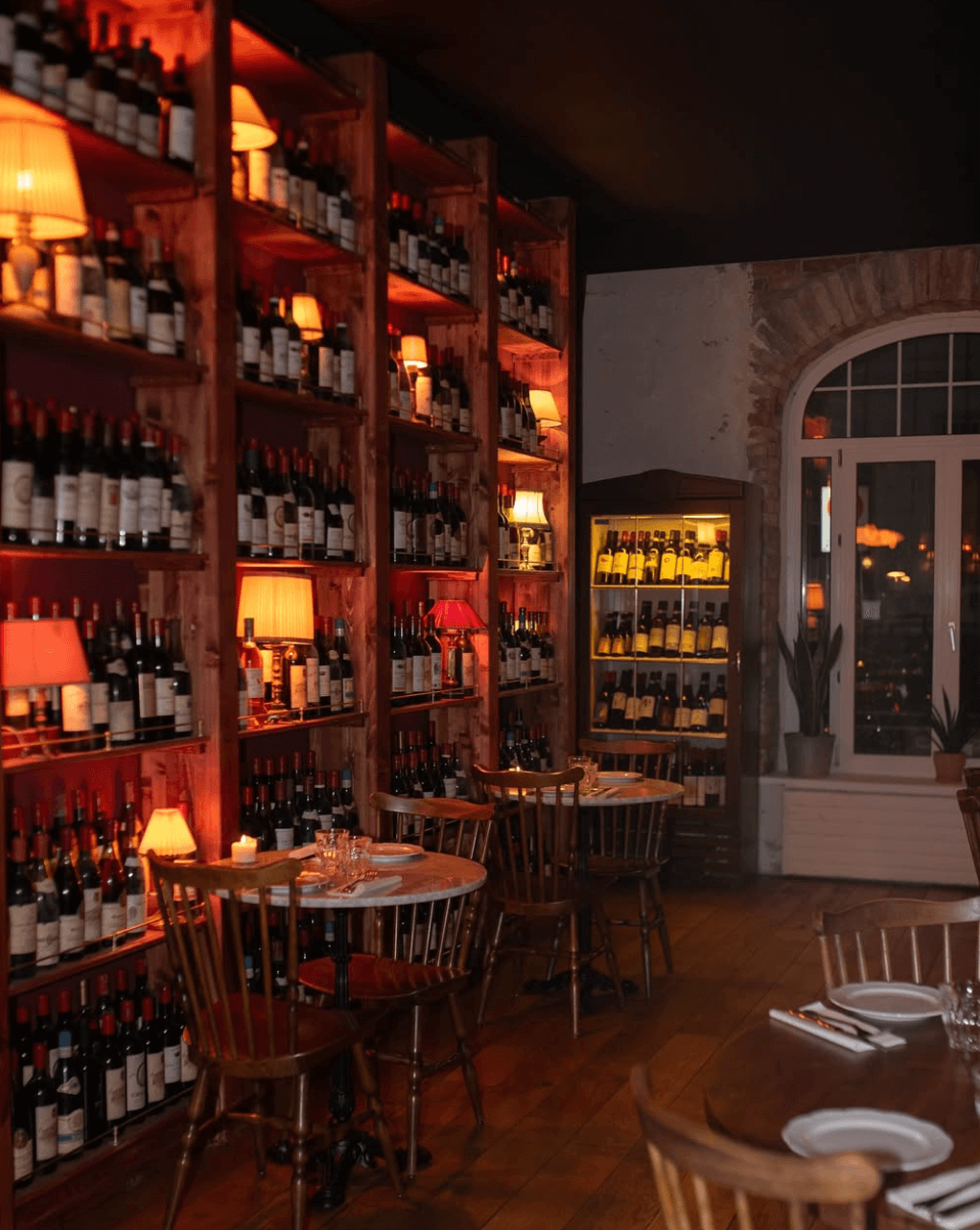 Une salle à manger chaleureuse avec des murs en briques et de grandes fenêtres, décorée de rayonnages remplis de bouteilles de vin, quelques lampes éclairant doucement la pièce, et des tables dressées avec des assiettes et des verres.