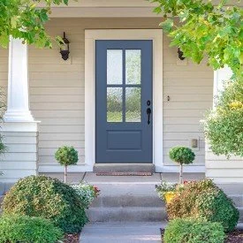 Front fiberglass door with blue color, flanked by green bushes and trees, with steps leading up to the entrance, part of a white house.