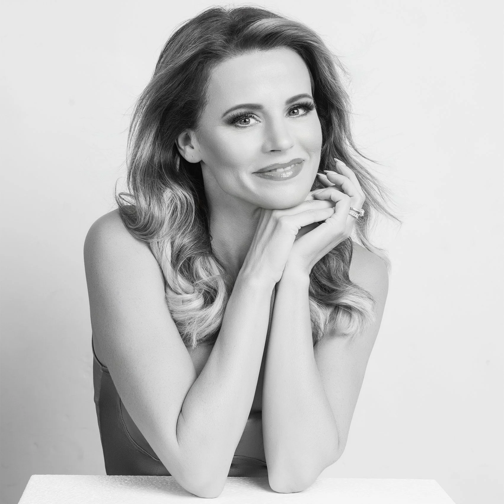 Black and white portrait of a woman with long, wavy hair, smiling softly, resting her chin on her hands, with a plain light background.