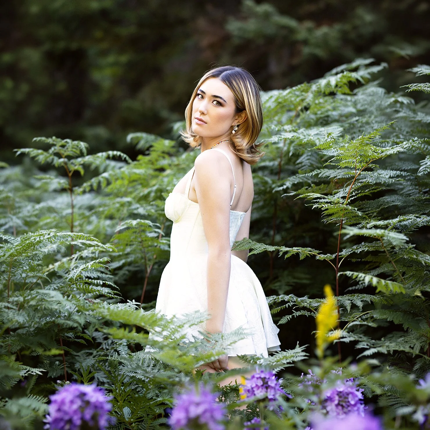 senior portraits near me at Gold Creek Pond by senior portrait photographer, Beauclair Photography of girl in white dress amongst ferns and purple flowers in foreground looking over her shoulder.