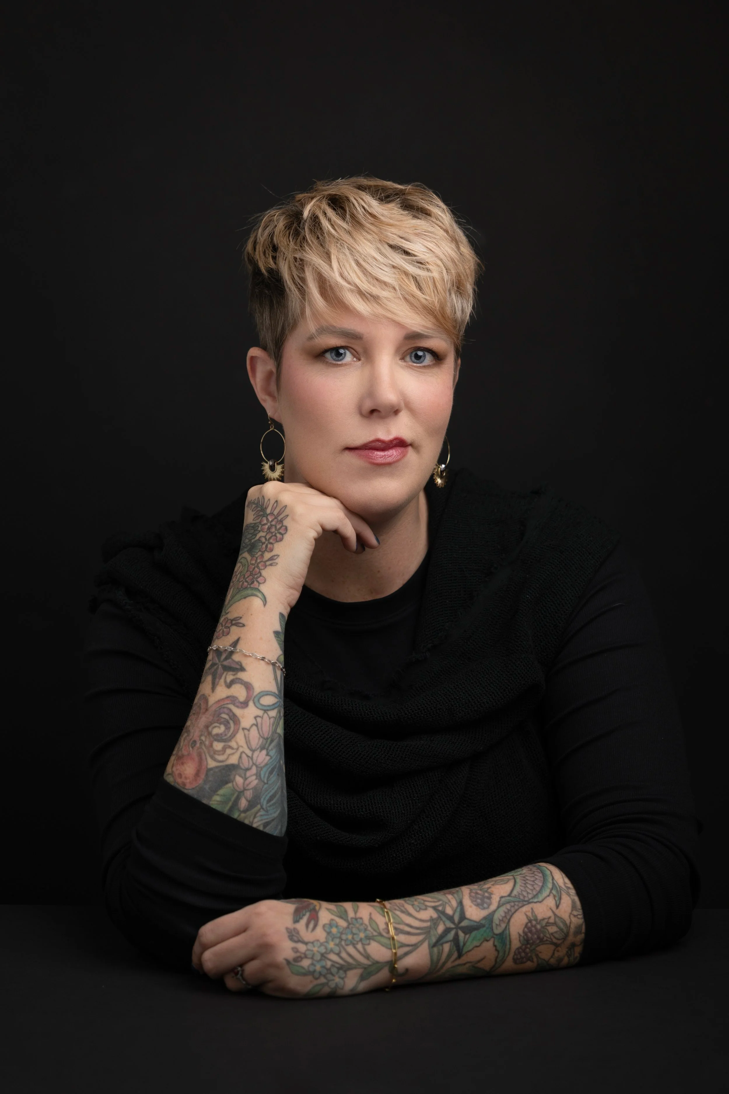 Portrait of a woman with short blonde hair, wearing earrings and a black top, sitting with her arm resting on a table, against a dark background.