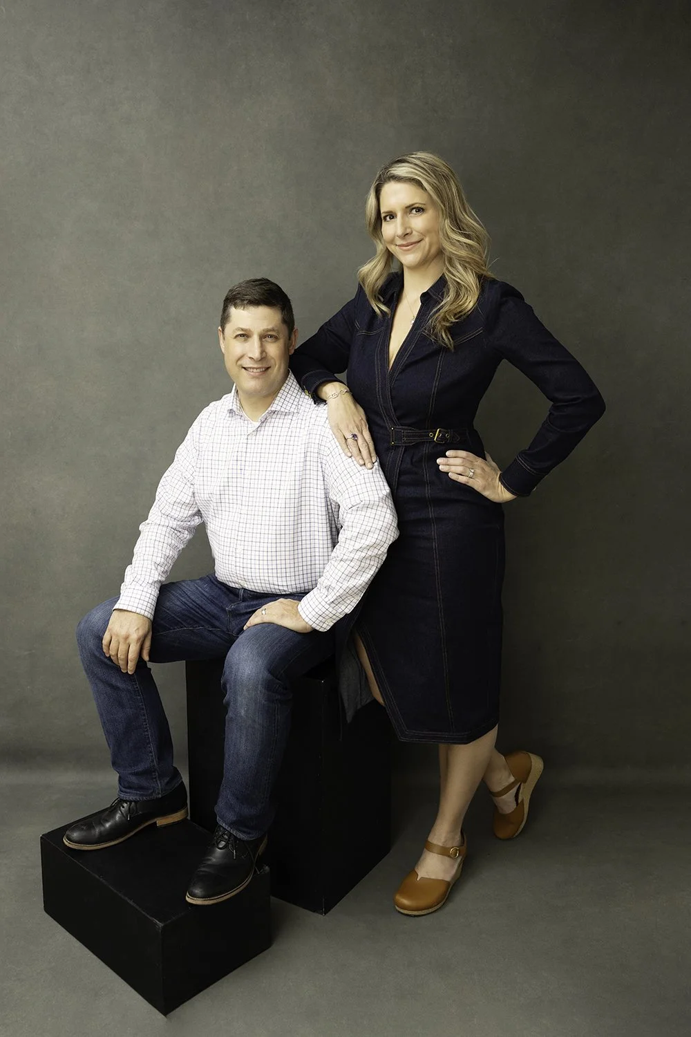 A man and woman pose together against a plain gray background. The man sits on a black cube, smiling, while the woman stands beside him with her hand on his shoulder, also smiling. The woman wears a dark denim dress with a slit, and tan high-heeled shoes. The man wears a white checked shirt, blue jeans, and black shoes.