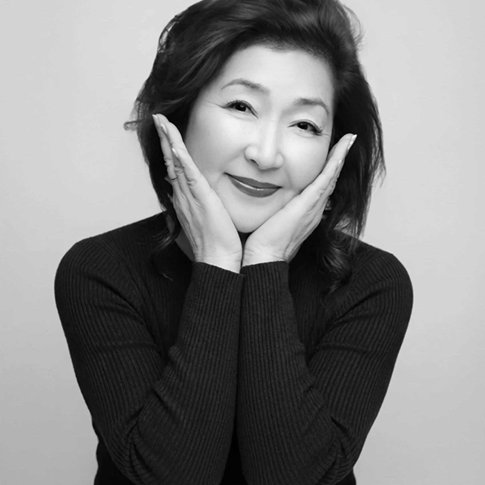 Black and white portrait of a smiling woman with her hands on her cheeks, wearing a dark ribbed long-sleeve top.