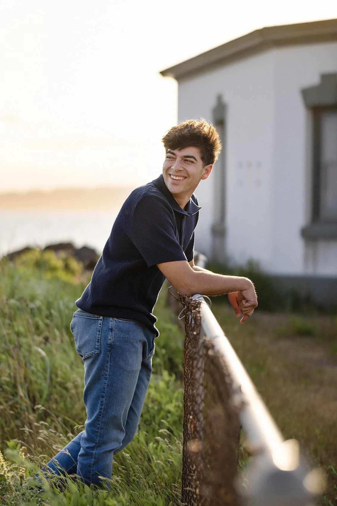 A young man with brown hair wearing a dark blue shirt and jeans leaning on a metal fence, outdoors during golden hour, smiling and looking to the side with a house and greenery in the background.