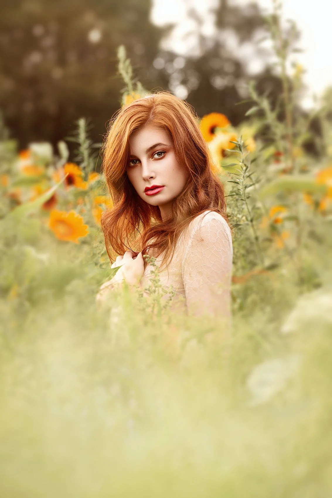 A young woman with red hair and red lipstick standing in a field of yellow flowers during sunset.