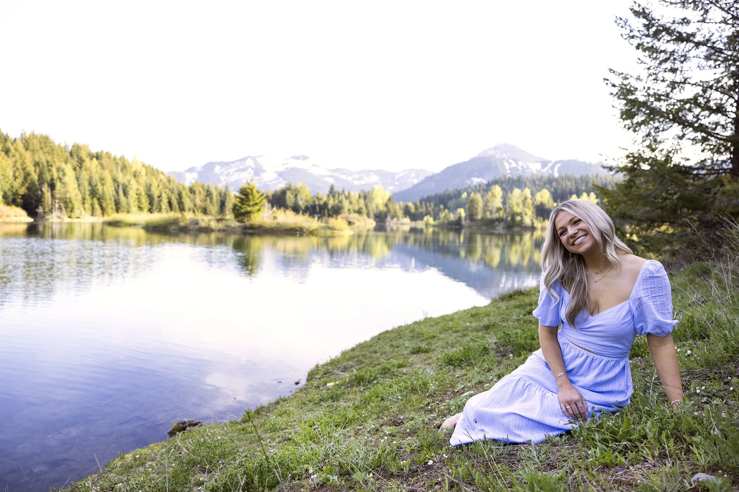 A young woman with long blonde hair sitting on green grass near a lake, smiling, with forested mountains and a bright sky in the background.