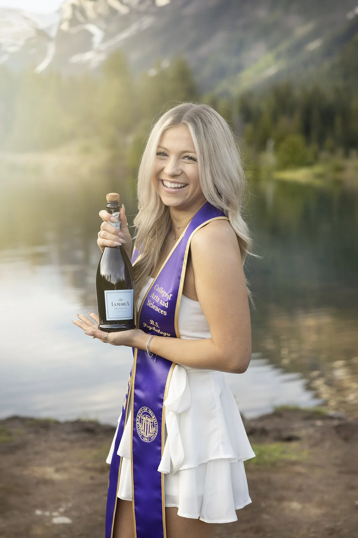 A young woman with blonde hair smiling and holding a bottle of sparkling wine in front of a lake with mountains in the background. She is wearing a white dress and a purple sash with gold text.