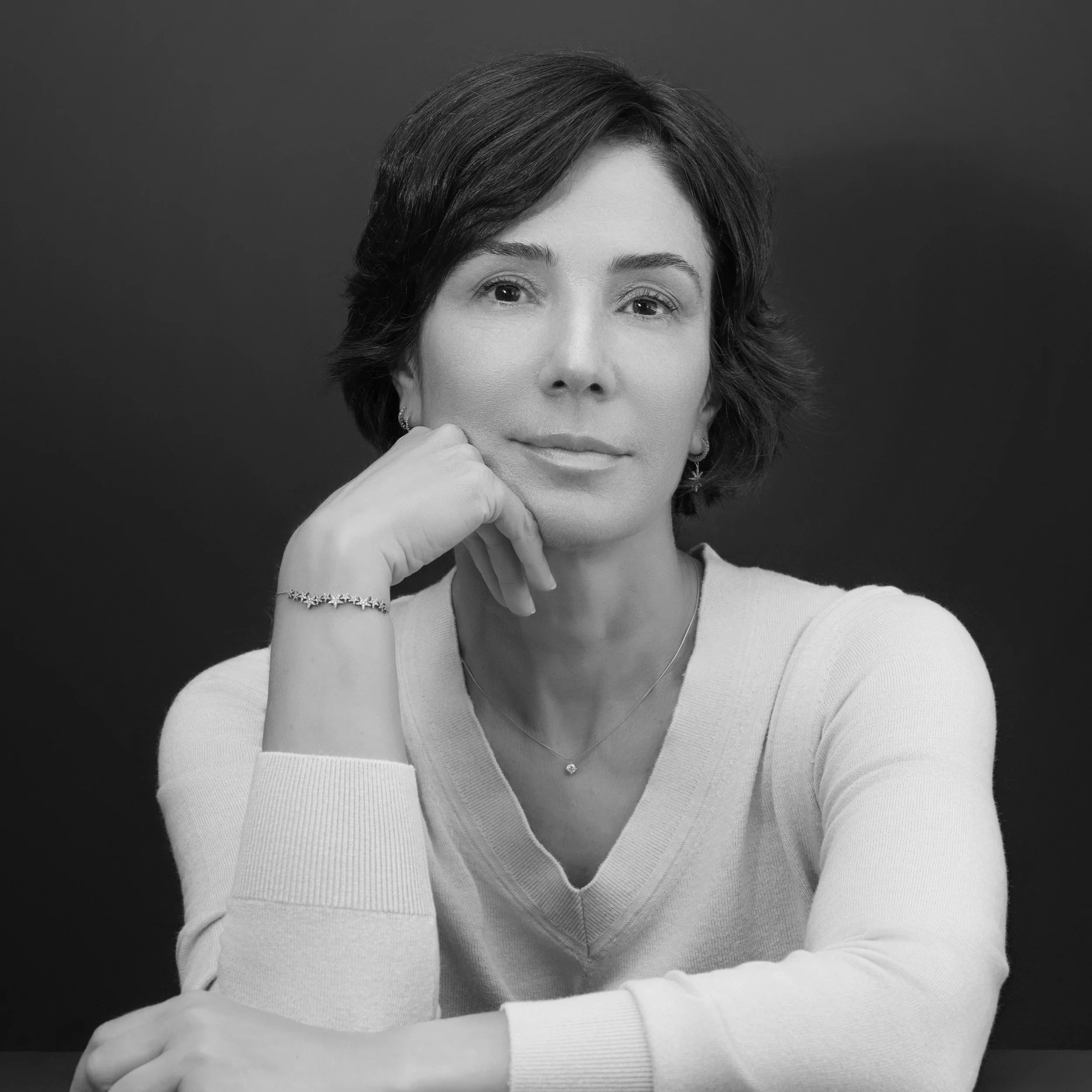 Black and white portrait of a woman with short hair, resting her chin on her hand, wearing a light-colored sweater, necklace, and earrings, with a neutral expression against a dark background.