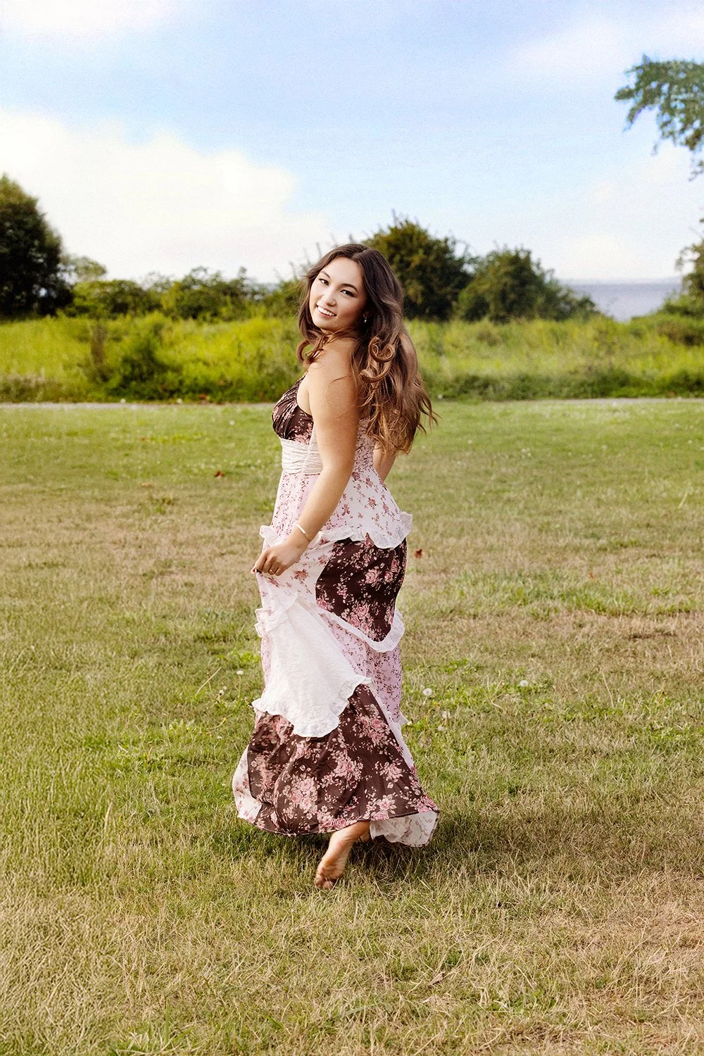 A young woman wearing a floral dress standing on grass in a park with trees in the background, smiling at the camera.