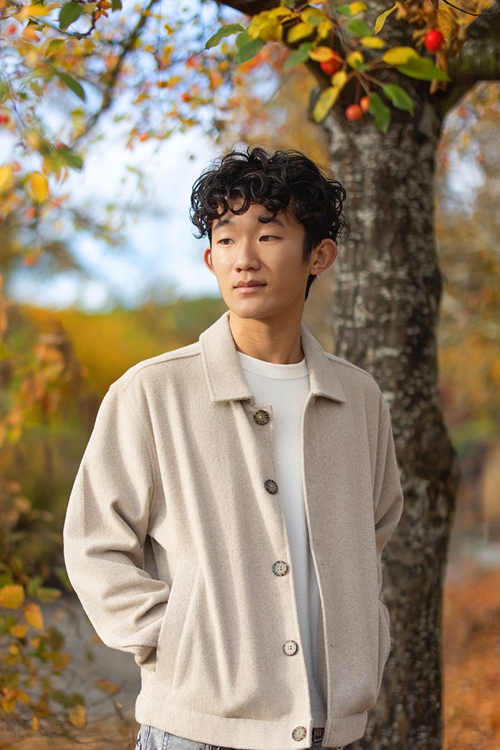 A young man with curly black hair stands outdoors during fall, wearing a beige coat over a white shirt, with fall-colored leaves and trees in the background.