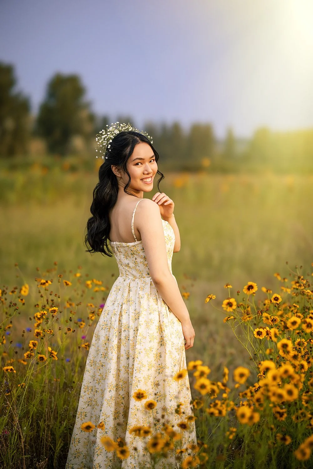 A woman in a floral dress with a flower crown, smiling in a field of yellow flowers at sunset during her senior portrait session with Beauclair Photography