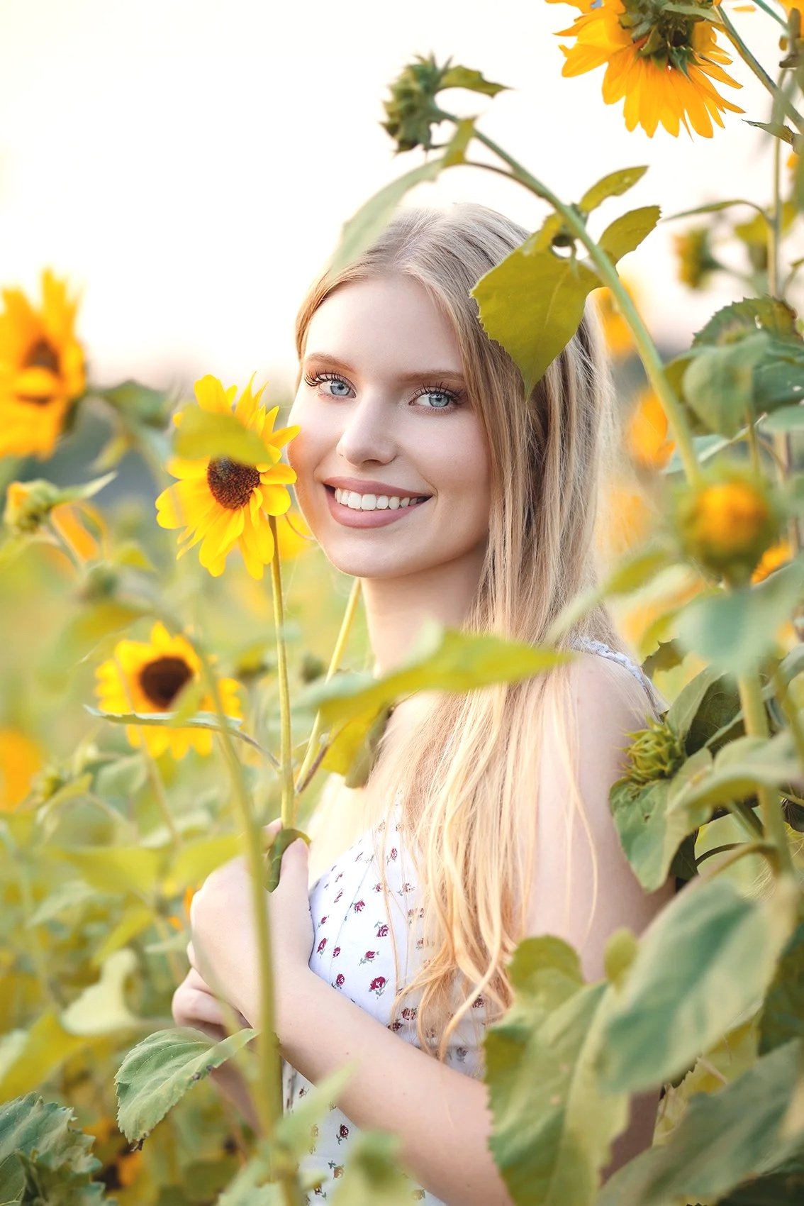 A young woman with long blonde hair and blue eyes smiling in a sunflower field during sunset during her senior portraits in Woodinville by Shannon Beauclair.