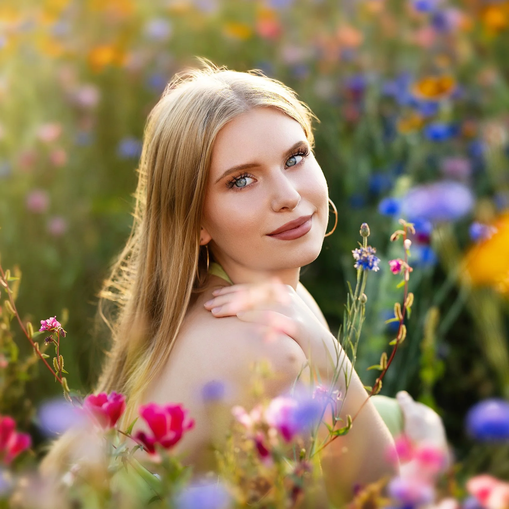 A young woman with blonde hair and blue eyes smiling in a field of colorful flowers.