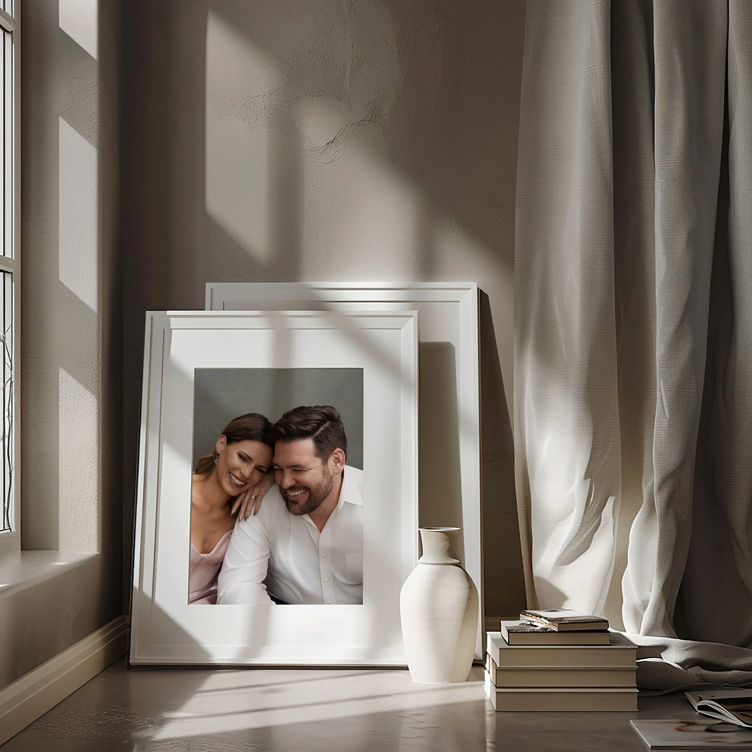 A framed photograph of a smiling woman and man leaning together, placed on the floor beside a white vase and a stack of books, with sunlight and curtains casting shadows on the wall.