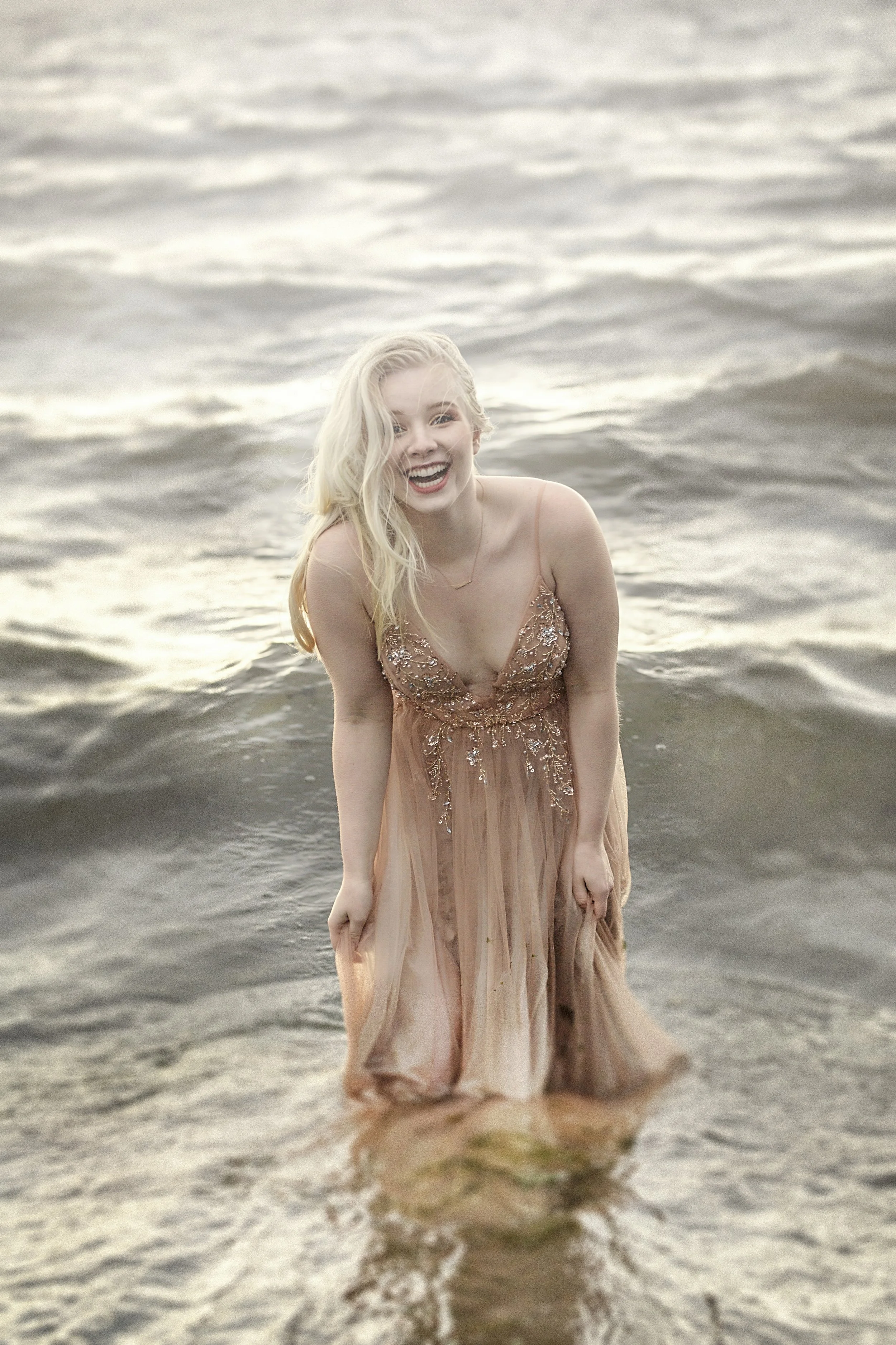 A woman with blonde hair in a peach-colored dress with embroidery, smiling and standing in the water at the beach during sunset.