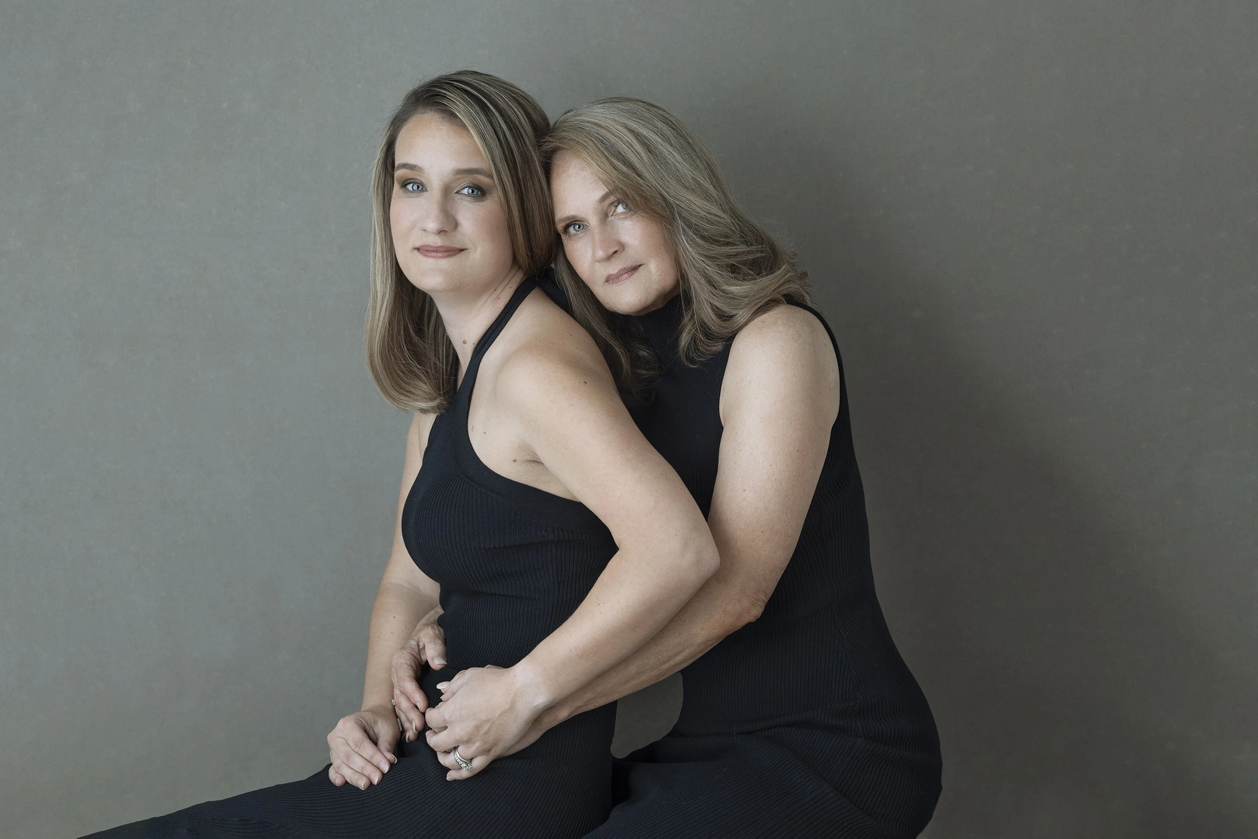 A woman with long blonde hair and a woman with shoulder-length gray hair sitting closely together, both wearing black sleeveless tops, against a neutral background.