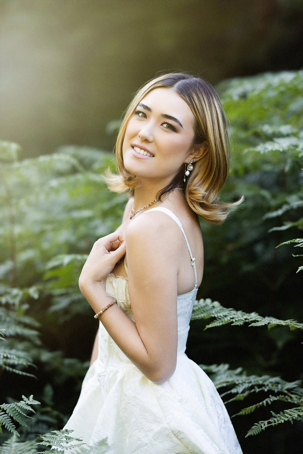 A young woman with shoulder-length hair, smiling and looking at the camera, wearing a white dress and jewelry, standing in a lush green outdoor setting.
