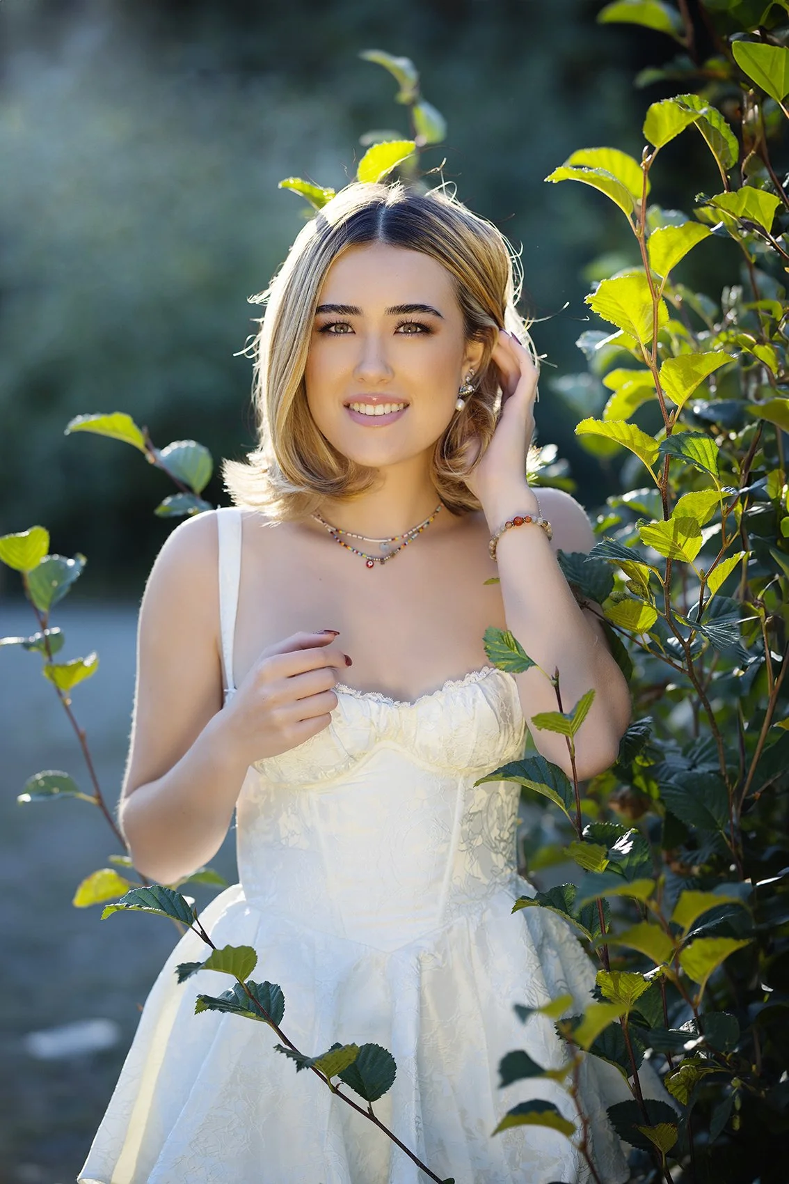A young woman with blonde hair and jewelry, wearing a white dress, standing outdoors near green foliage.