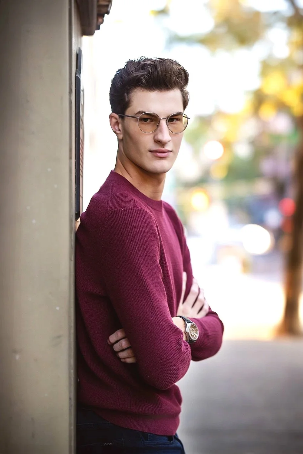 A young man with dark hair wearing glasses and a red sweater with his arms crossed, standing outdoors and leaning against a wall. Blurry trees and lights are in the background in Pioneer Square for urban senior portraits.
