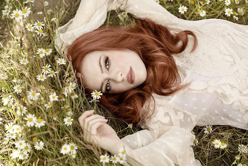 A woman with red hair lying in a field of white daisies, wearing a white lace dress.