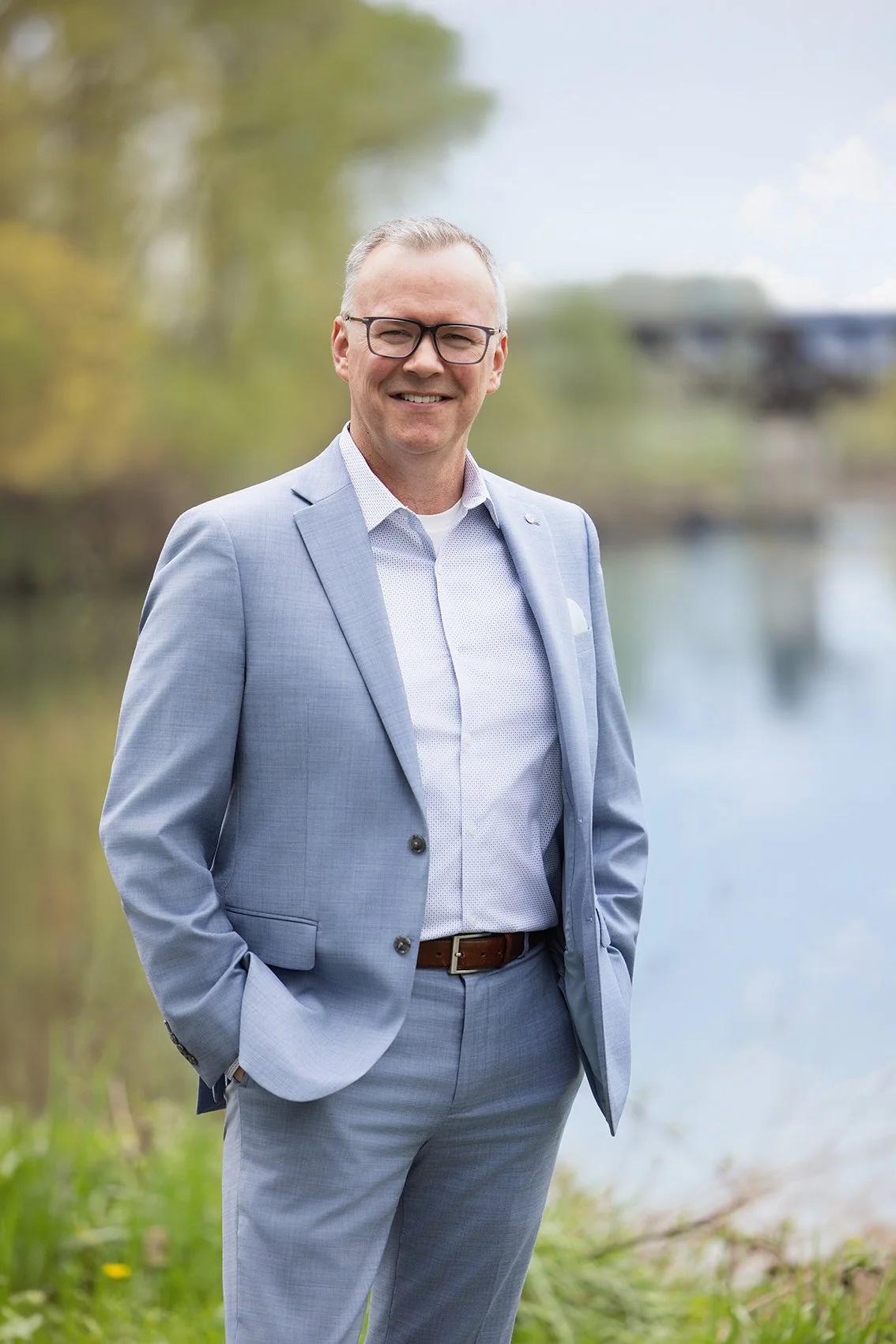 A man in a light gray suit stands outdoors by a river, smiling at the camera, with a background of trees and water.