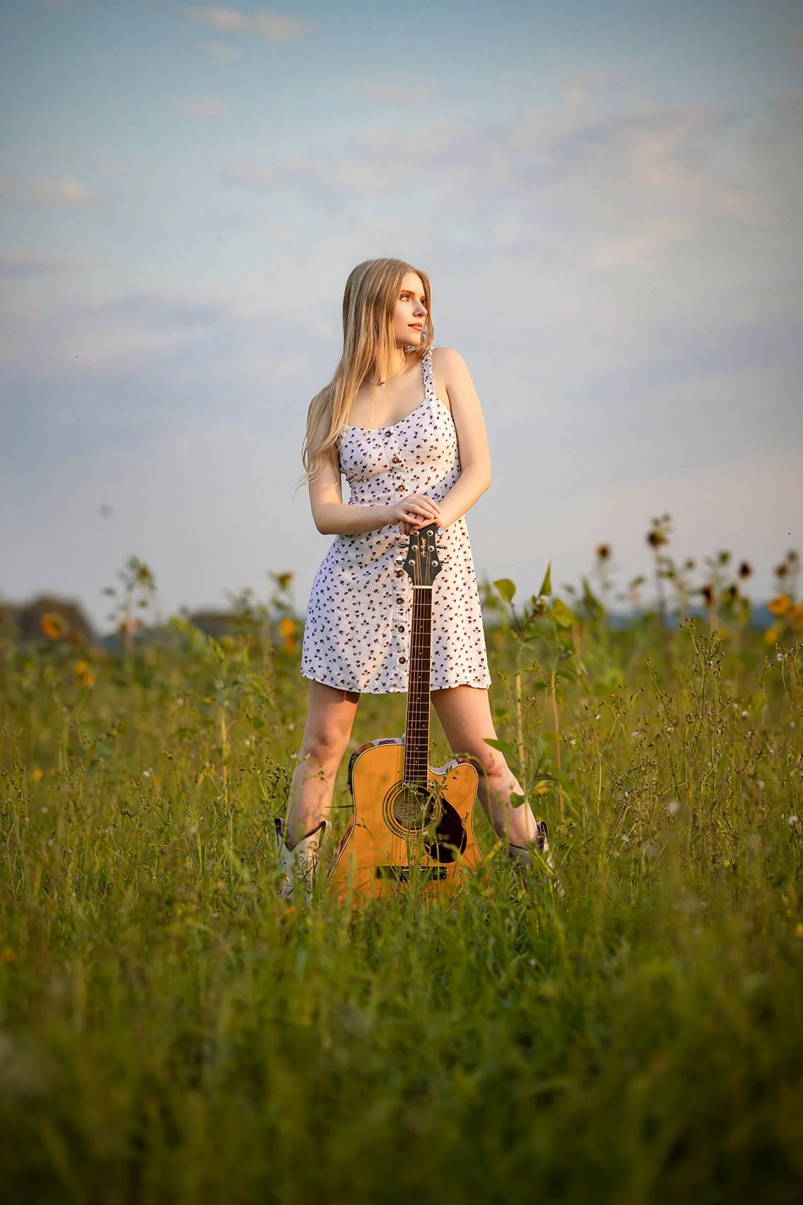 A young woman in a summer dress holding a guitar in a field of tall grass and wildflowers, with a partly cloudy sky overhead.