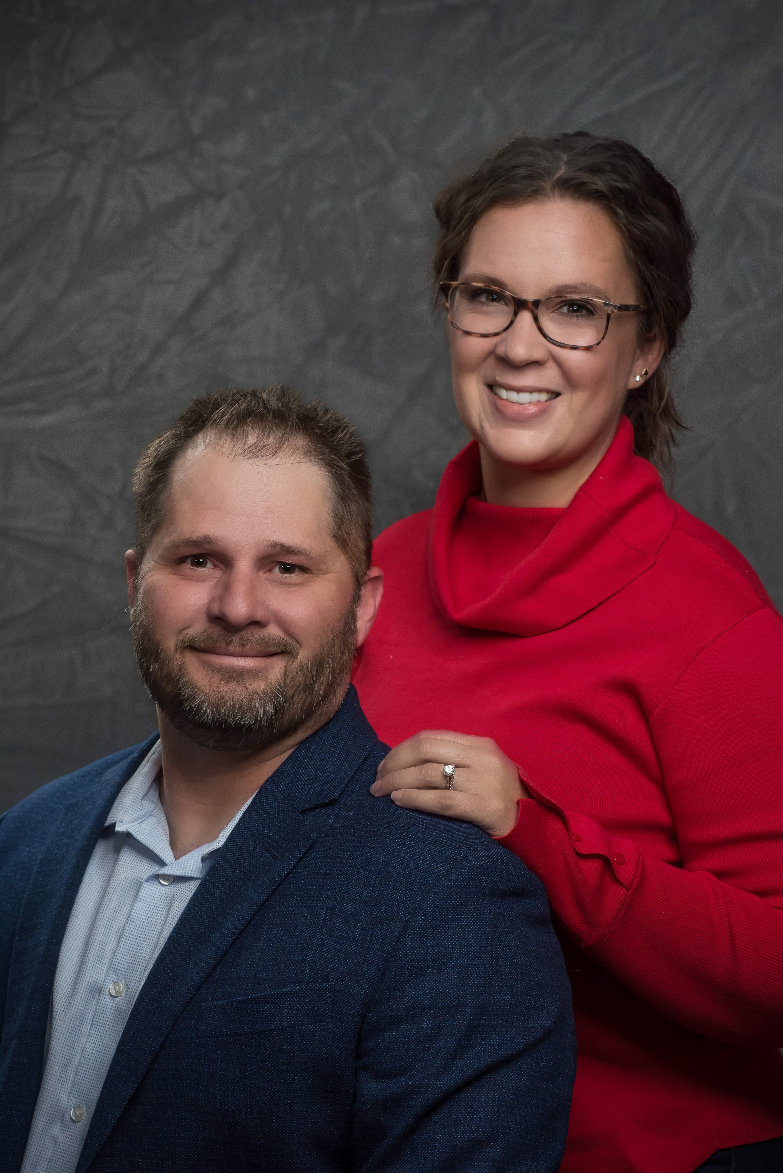 A couple posing for a portrait, with the man wearing a blue jacket and the woman in a red sweater, her hand resting on his shoulder.