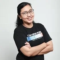 A woman with dark hair and glasses smiling with arms crossed, wearing a black T-shirt, standing against a light background.