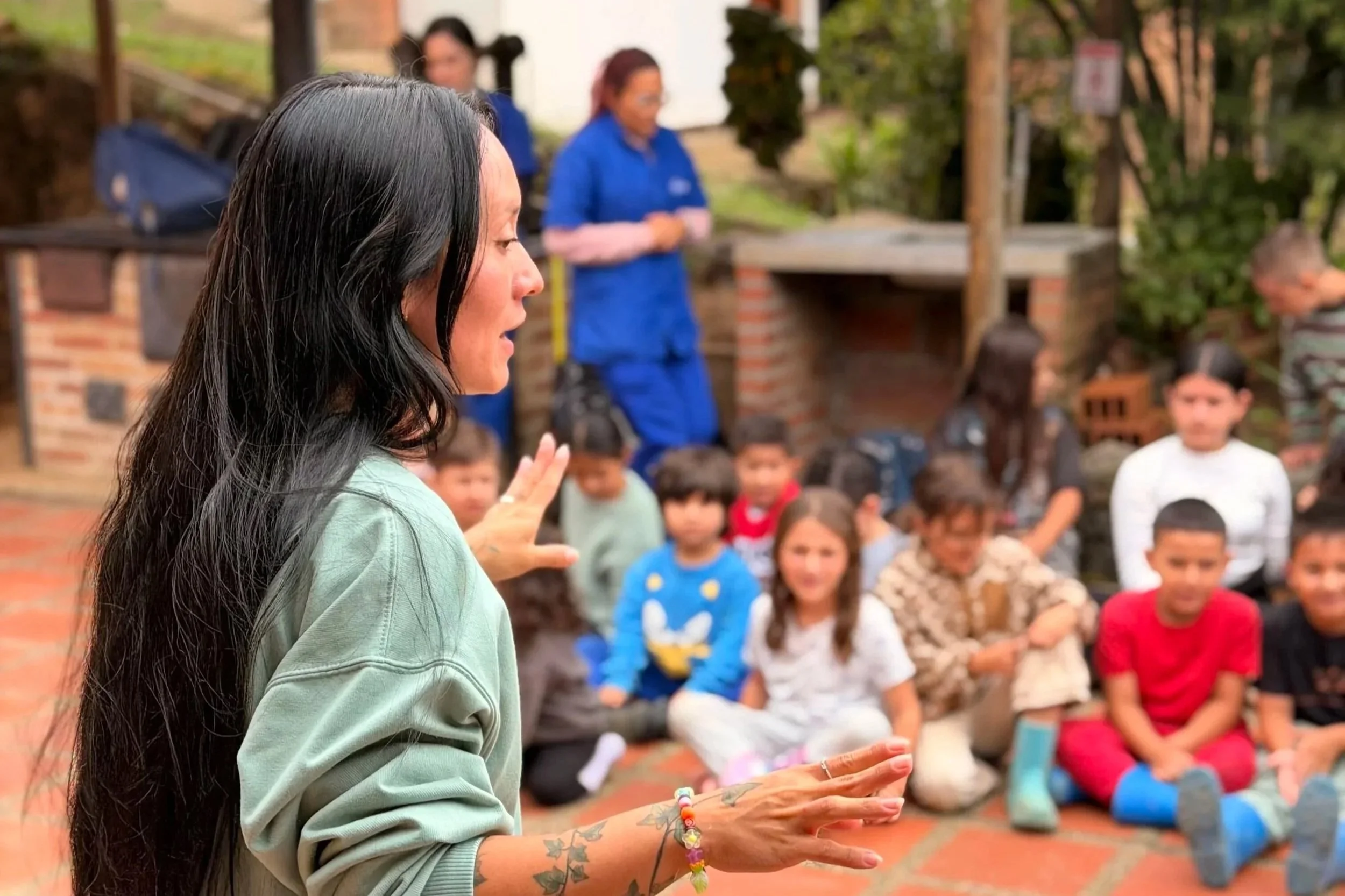 An instructor sitting on the ground outdoors, gesturing with her hands while speaking to a group of young school children seated in a circle in front of a brick building.
