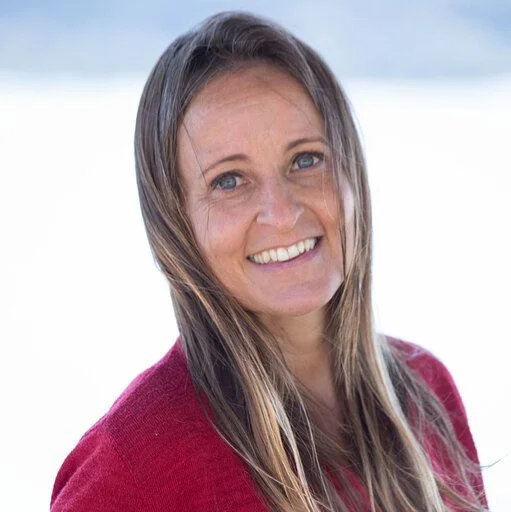 A woman with long brown hair and blue eyes, smiling outdoors with a blurred natural background, wearing a red sweater.