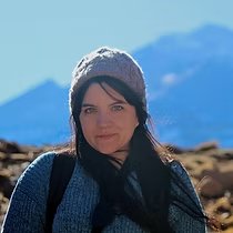 Woman with long dark hair wearing a gray knit beanie and blue sweater, standing outdoors with mountain scenery in the background.