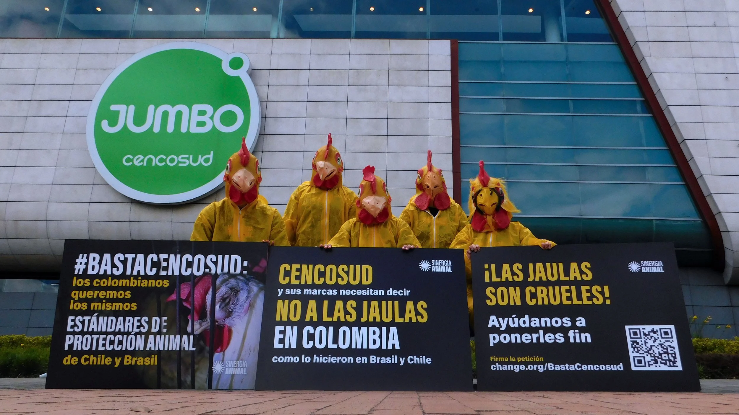 Five Sinergia Animal activists wearing chicken costumes and masks hold protest banners against battery cages in front of a Jumbo Cencosud supermarket in Colombia.