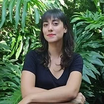 A woman with dark hair and bangs, wearing a black shirt, standing in front of lush green plants.
