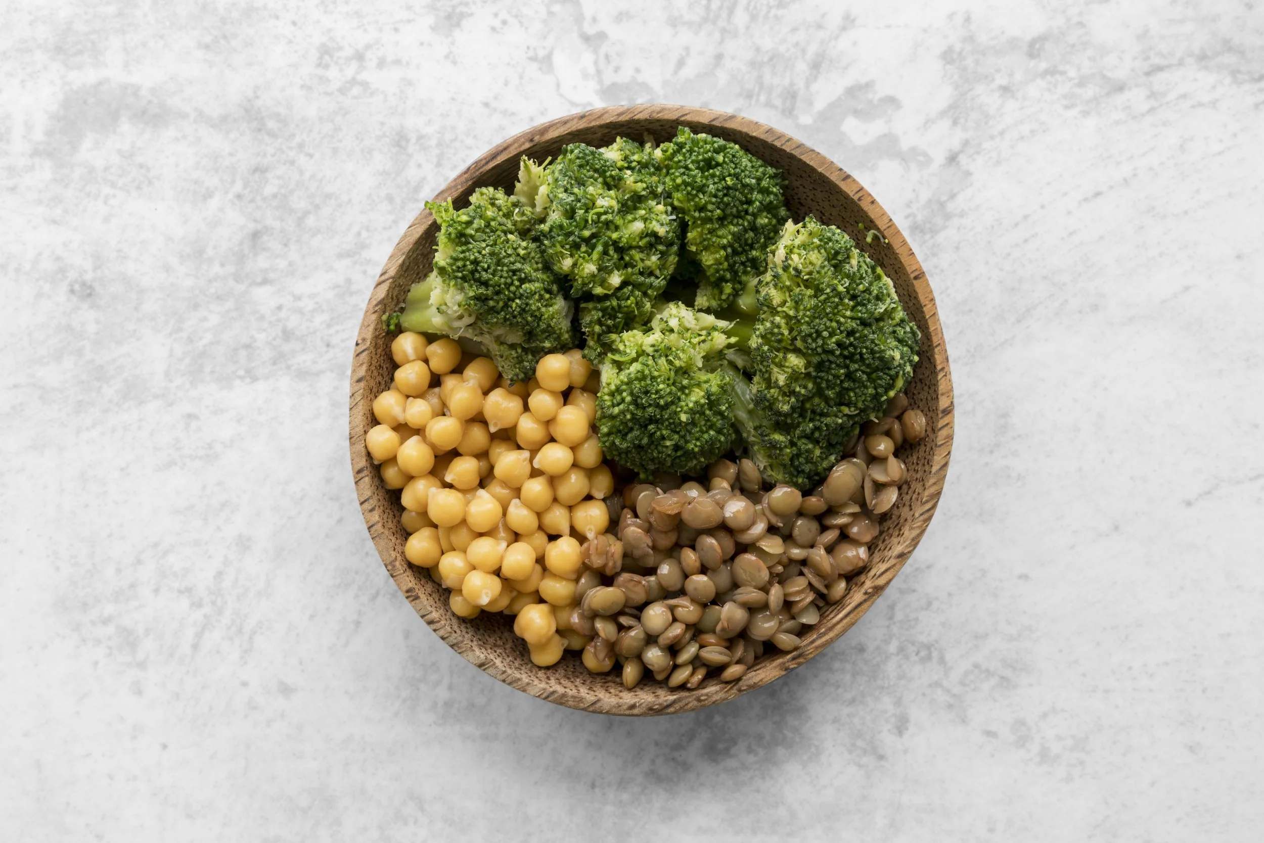 A top-down circular shot with a black background showing a wooden bowl filled with vibrant green steamed broccoli florets, cooked chickpeas, and brown lentils arranged in sections