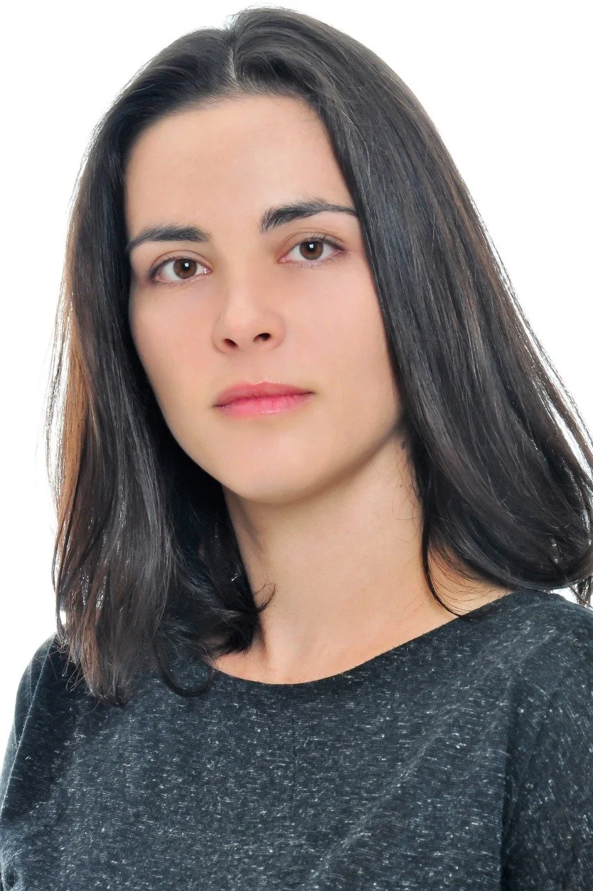A woman with straight dark brown hair wearing a dark gray textured shirt, looking directly at the camera with a neutral expression, against a plain white background.