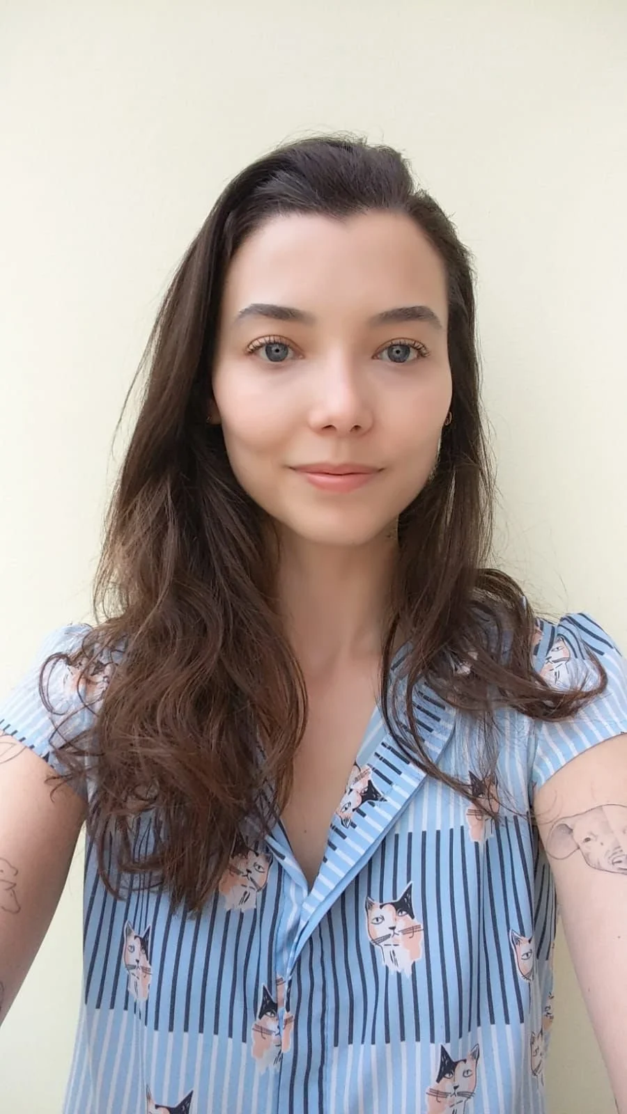 A woman with long brown wavy hair and blue eyes taking a selfie against a plain light-colored background, wearing a blue striped shirt with cat faces.