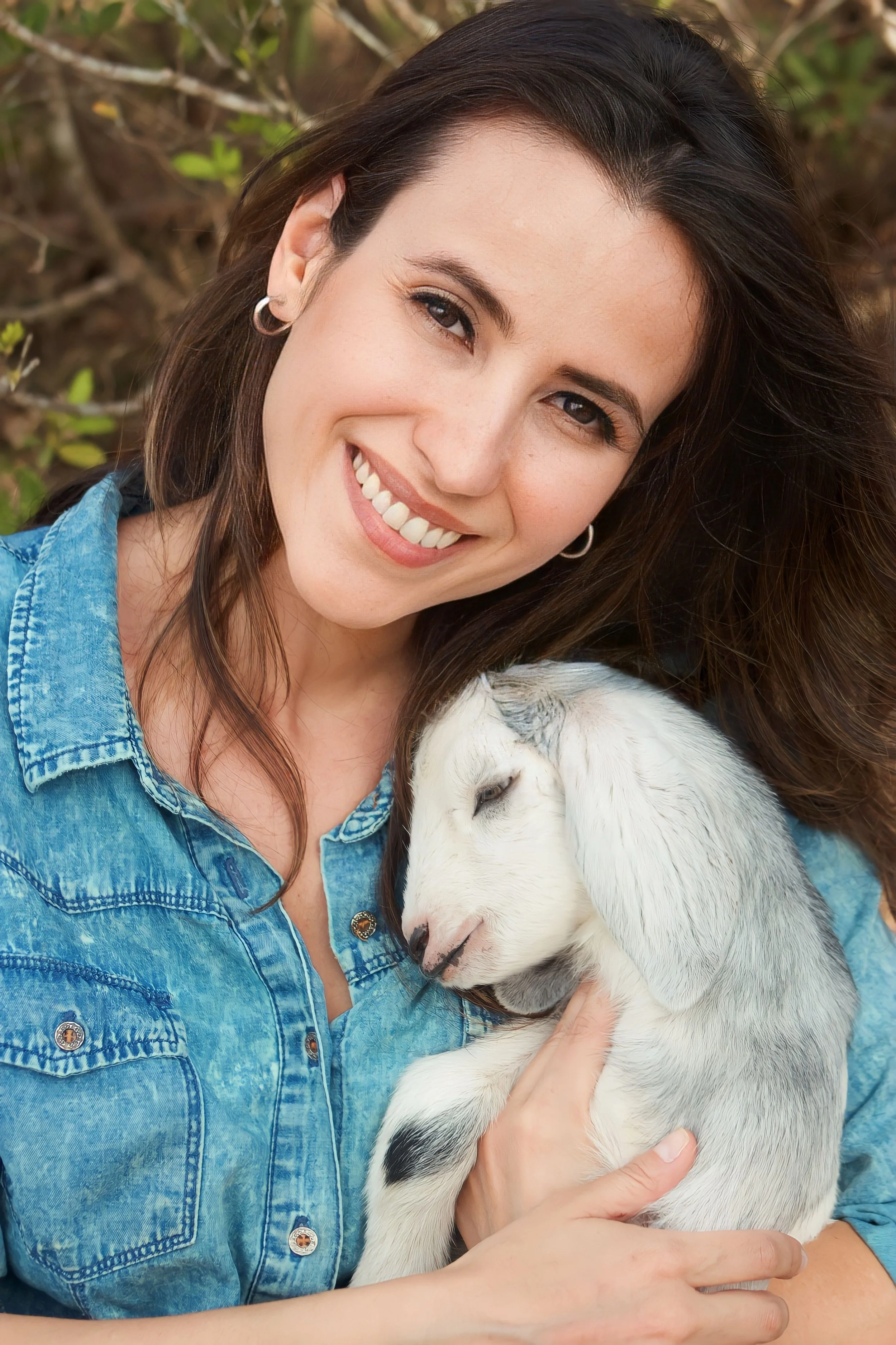 A woman with tousled dark hair, wearing a blue denim shirt, is smiling and holding a white puppy with black spots close to her chest, outdoors with green foliage in the background.