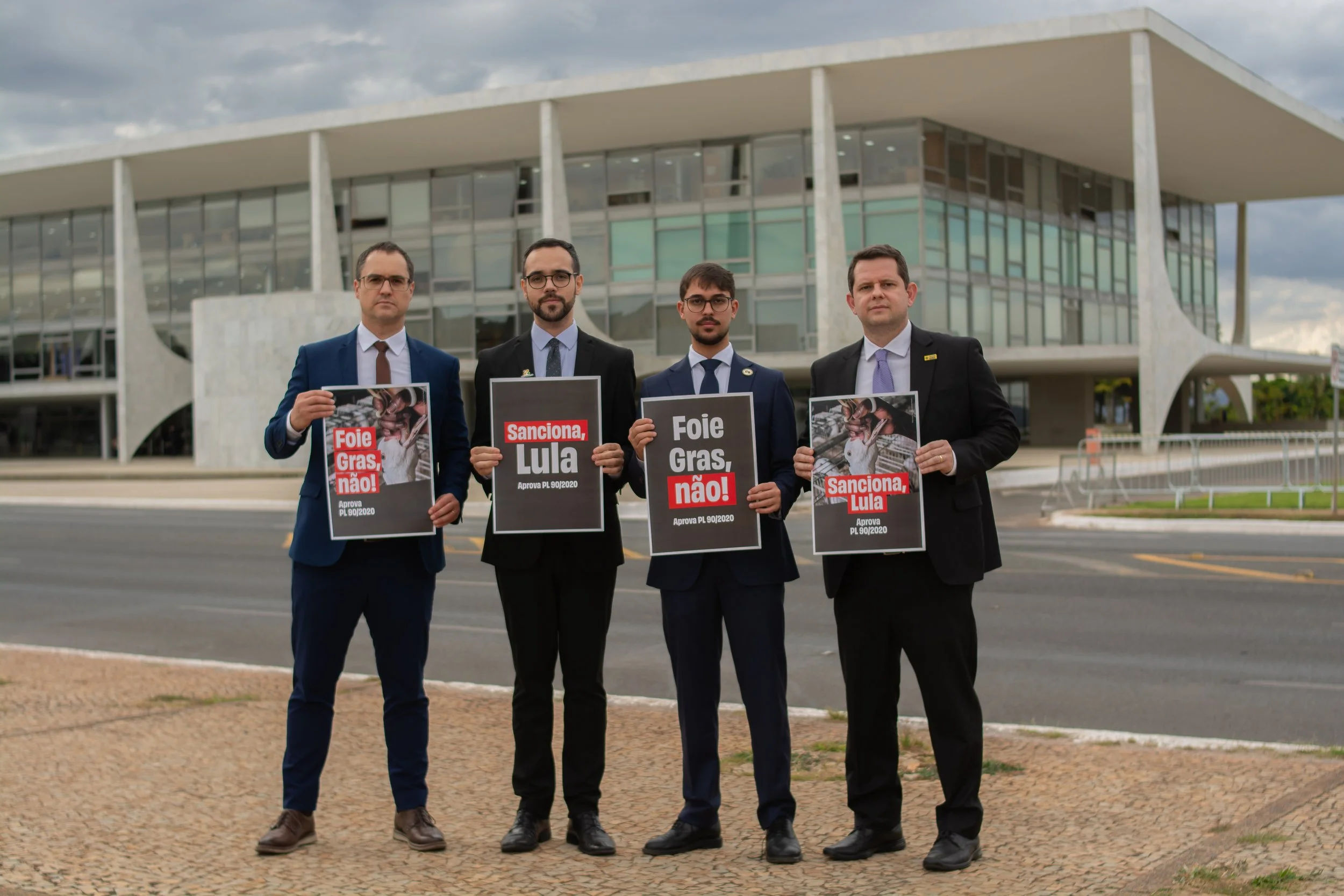 Four men in business suits standing outdoors in front of a government building in Brasília, holding signs advocating against foie gras with messages reading "Foie Gras, Não!" and "Sanciona, Lula".