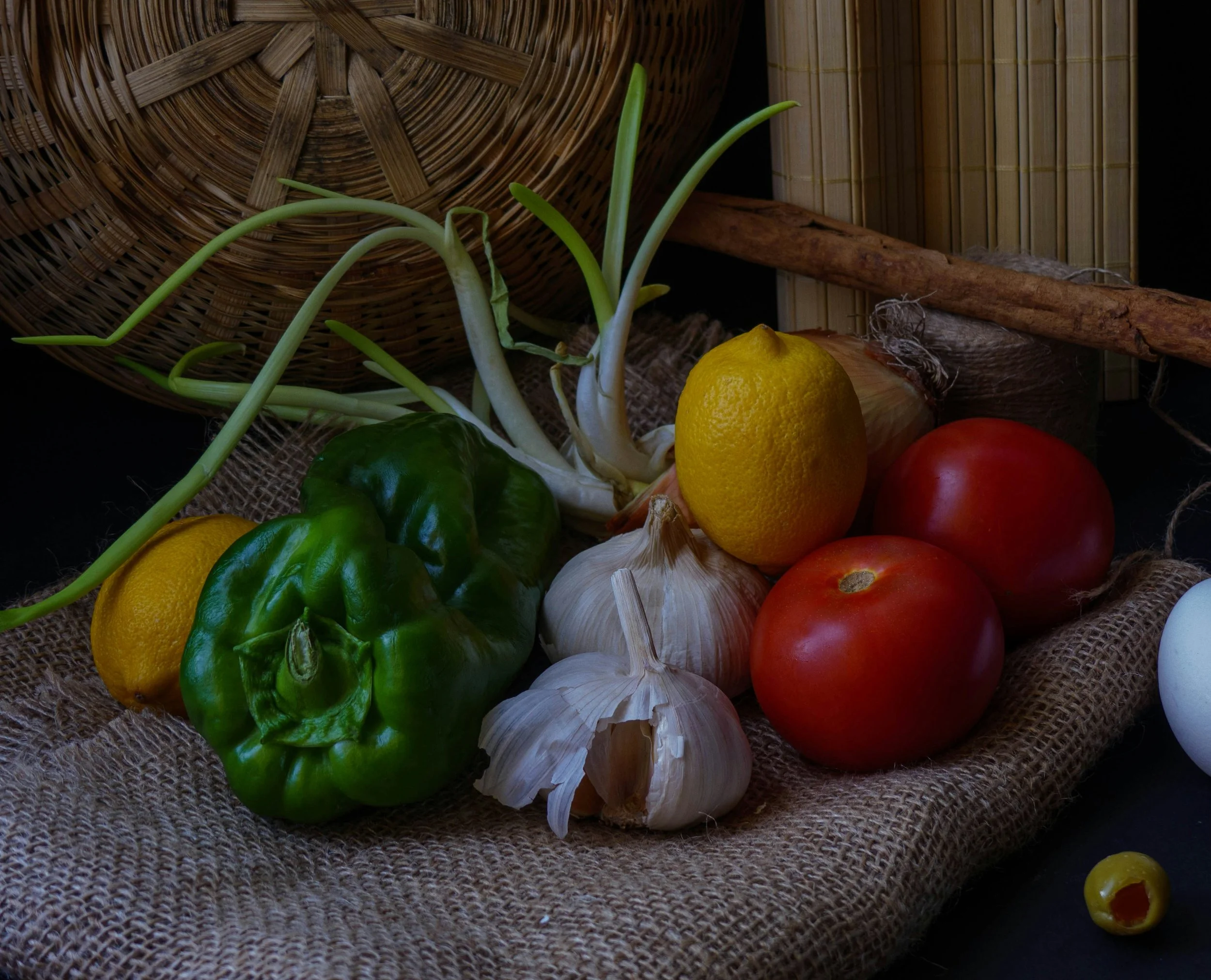 Various fresh vegetables including lemons, garlic, green bell pepper, tomatoes on burlap fabric with woven baskets in the background.