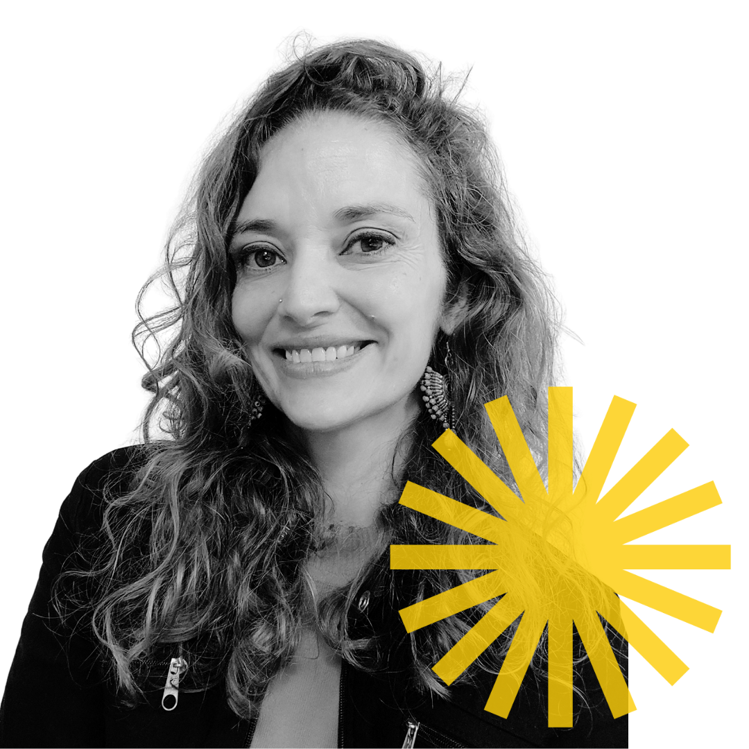Black and white portrait of a woman with curly hair, wearing earrings and a jacket, smiling at the camera, with a yellow radial burst graphic overlapping the bottom right corner.