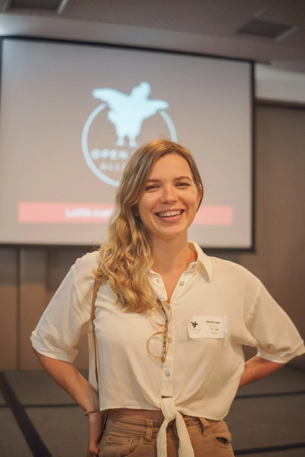 A smiling woman with wavy blonde hair wearing a beige tied-up shirt stands in front of a screen with a logo that features a bird, possibly an eagle, and text that is blurred. She has glasses hanging from her shirt and a name tag on her chest.