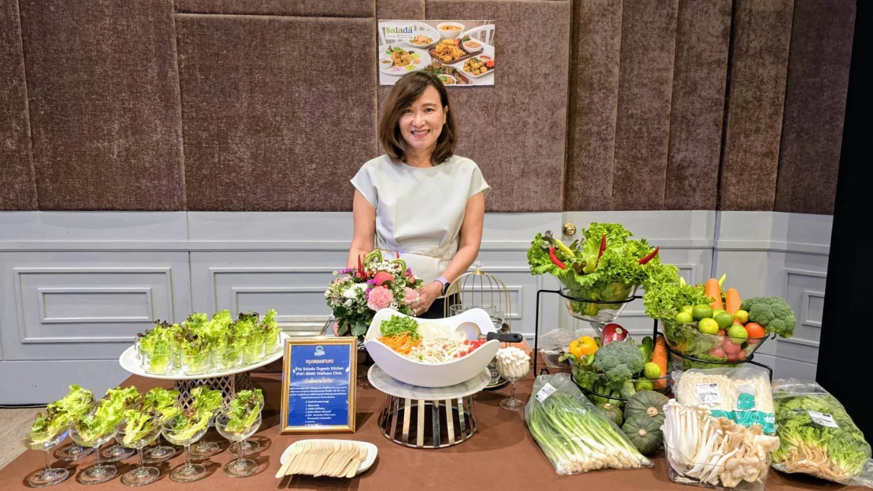 Benjawan Charuchiewpanich, owner of Salada Organic Kitchen, smiling behind a display table filled with fresh vegetables, salads, and floral arrangements against a textured wall backdrop.