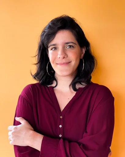 A woman with shoulder-length dark hair, wearing hoop earrings and a burgundy blouse, smiling and posed against a yellow background.