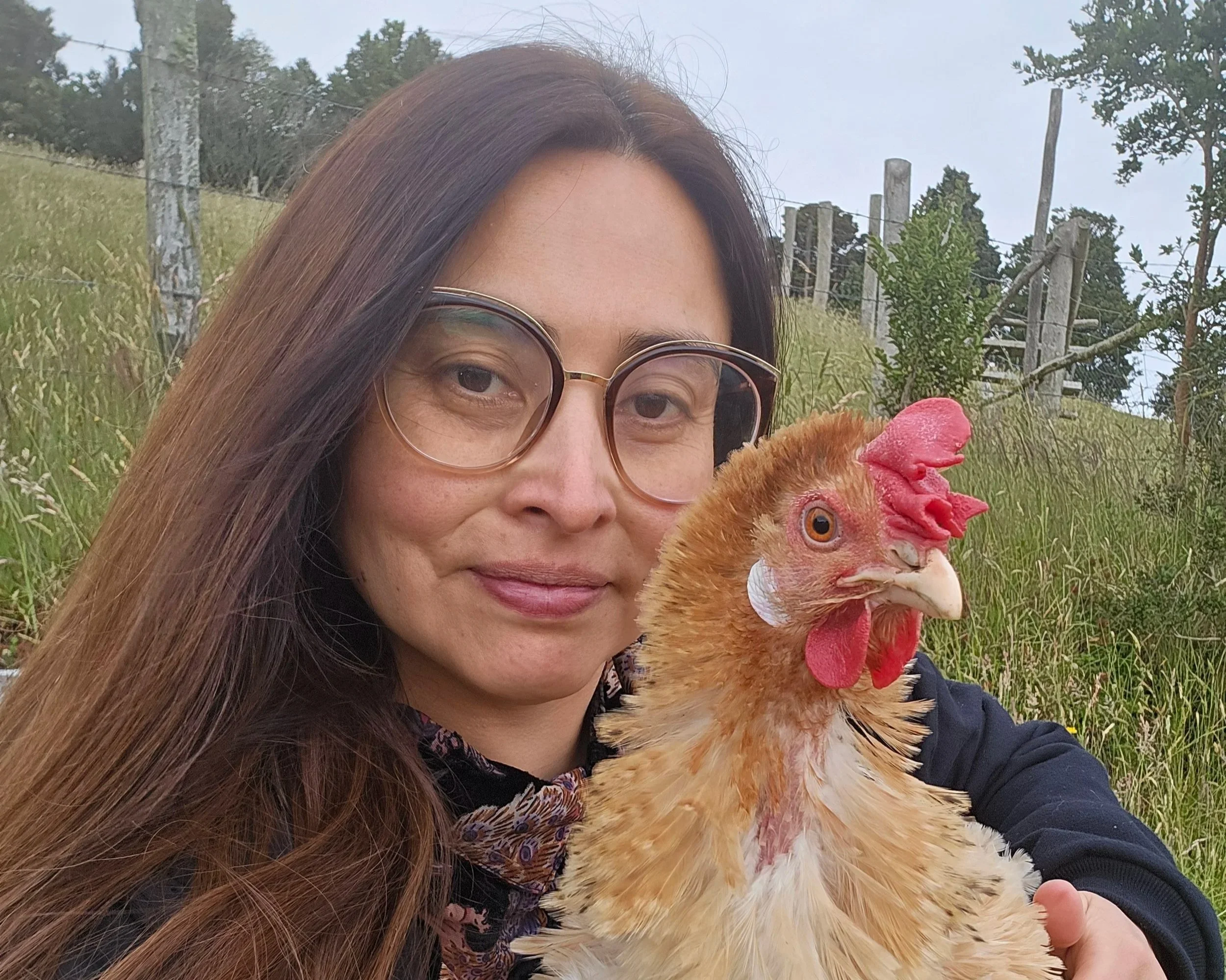 A close-up portrait of a woman with long brown hair and glasses smiling gently outdoors next to Elena, a rescued brown laying hen with a bright red comb, on a grassy hillside.