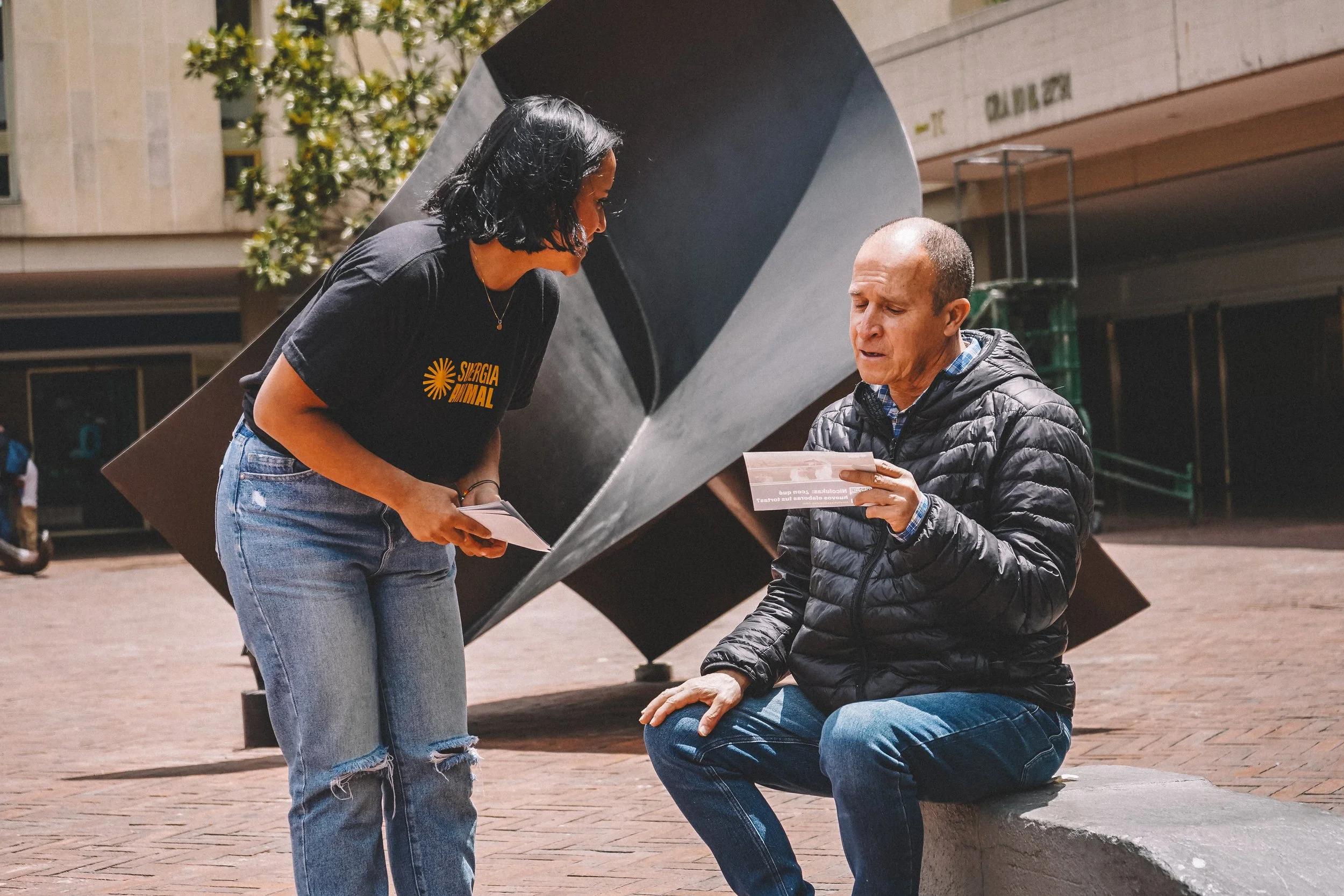 A woman with shoulder-length black hair, wearing a black t-shirt and ripped blue jeans, is standing and talking to a man with short hair, wearing a black jacket and blue jeans. The man is sitting on a stone bench and holding a piece of paper. They are outdoors, near a large abstract metal sculpture, surrounded by a city setting with a building and trees in the background.