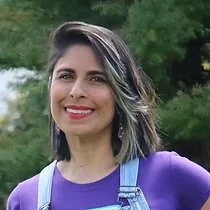 A woman smiling outdoors with green foliage in the background, wearing a purple top and denim overalls.