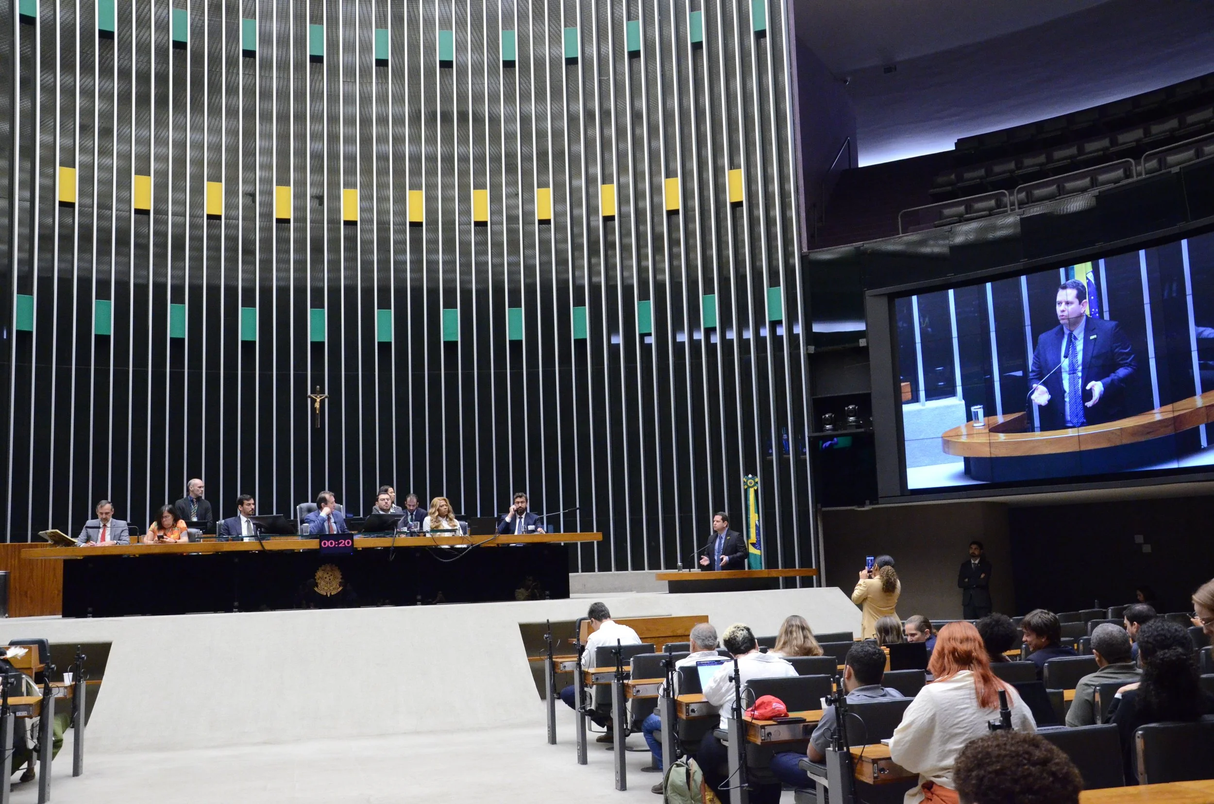 Arthur Regis speaking at a podium during a Special Session in Brazil's Chamber of Deputies, displayed on a large screen above seated lawmakers and attendees in the assembly hall.