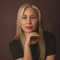 Portrait of a woman with blonde hair, wearing a black top, resting her chin on her hand against a brown background