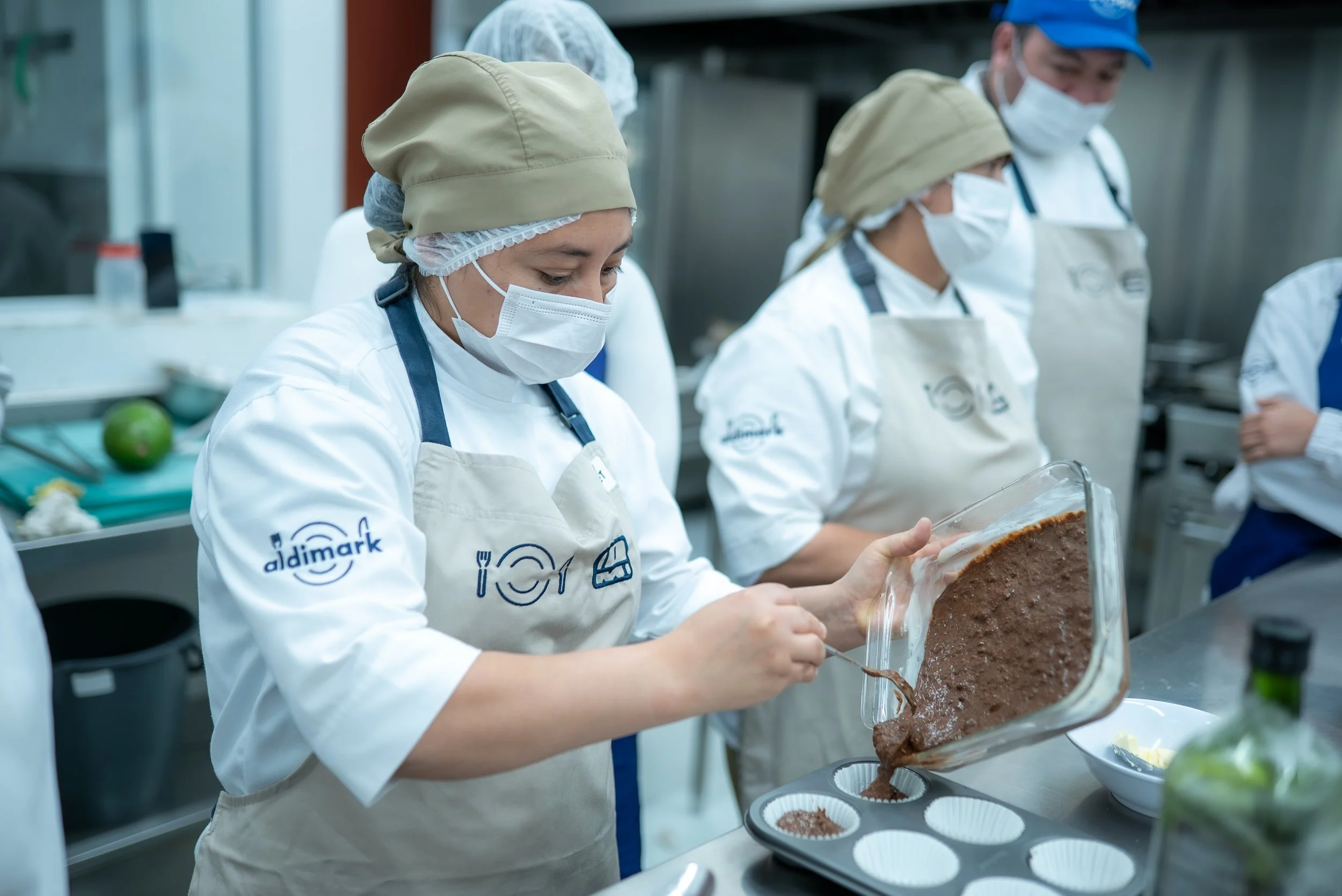 A kitchen worker wearing a chef uniform, apron, face mask, and hairnet carefully pouring a batter mixture into a muffin pan in a commercial kitchen setting.