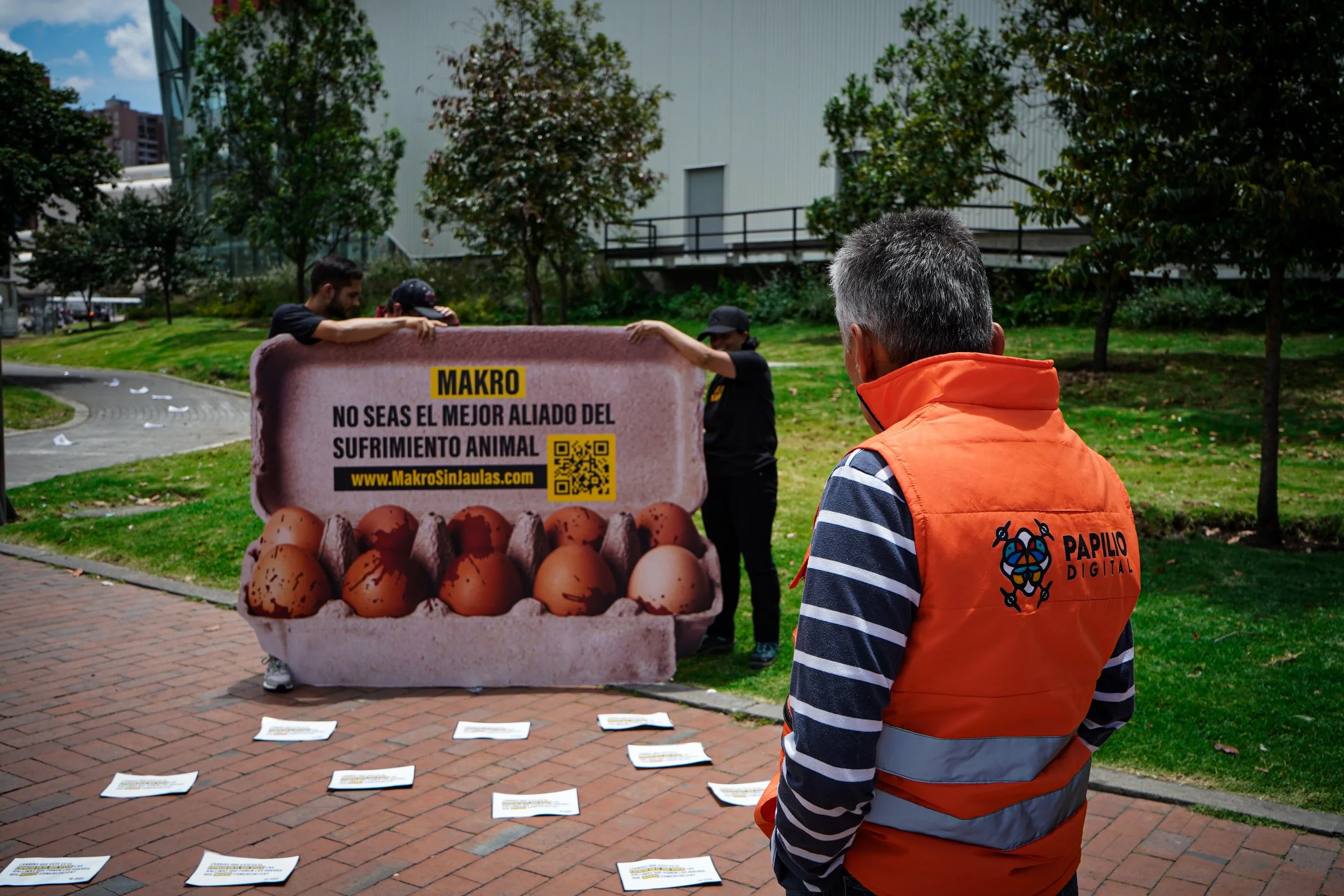 A person wearing an orange vest with the logo 'Papilo Digital' is standing outdoors facing a display about animal suffering and eggs. There are two other people on the other side of the display, examining it. The display features a large image of eggs and a QR code, with text in Spanish that reads: 'Makro, No seas el mejor aliado del sufrimiento animal'.